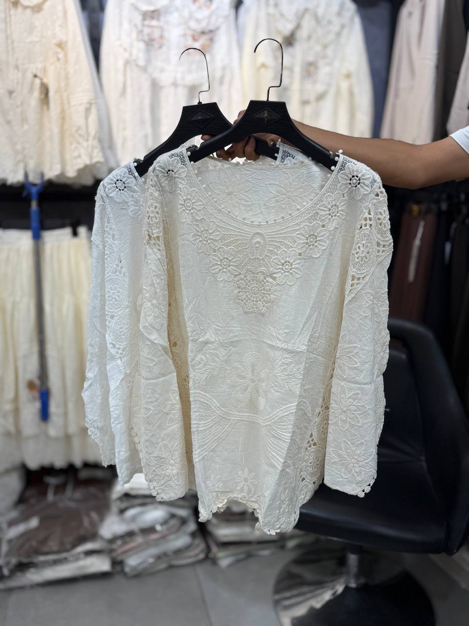 بلوز شومیز گیپوری سفید نخی با گلدوزی گلهای برجسته و چیندار، یقه گرد مروارید دوزی، آستین بلند و لبههای توری، نمای جلو و پشت محصول روی چوب لباسی مشکی در فروشگاه.